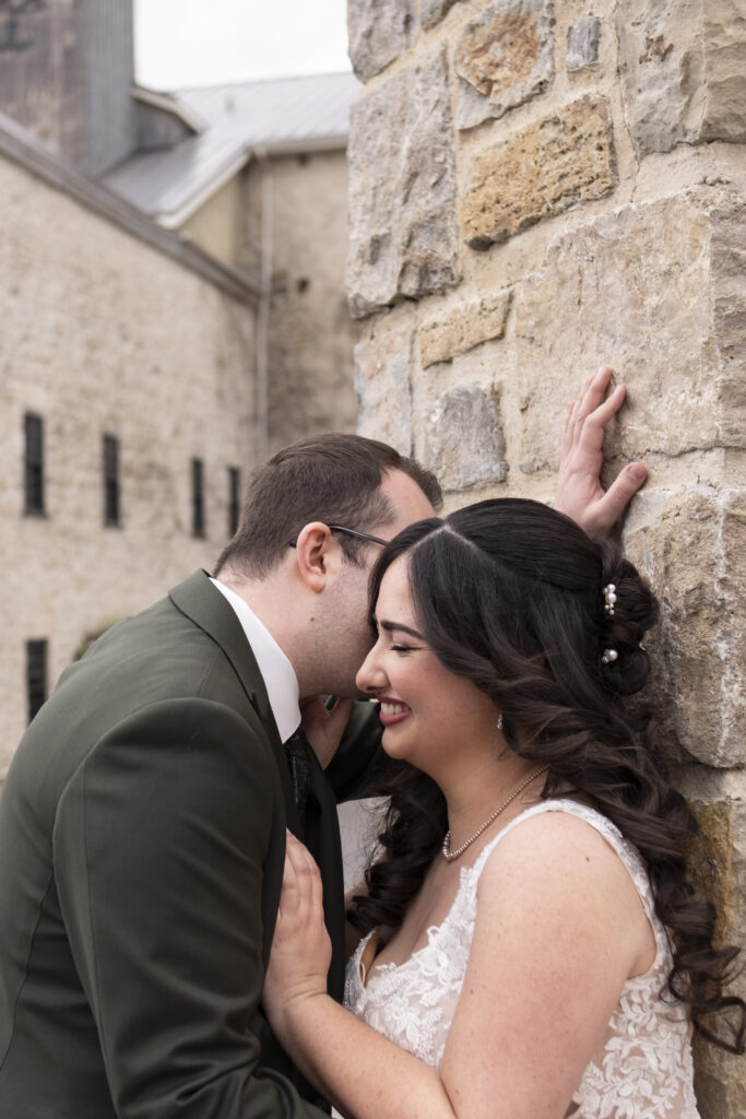 The bride smiles while the groom leans into her ear while standing at the stone gatepost at the Elora Mill, in Elora Ontario.
