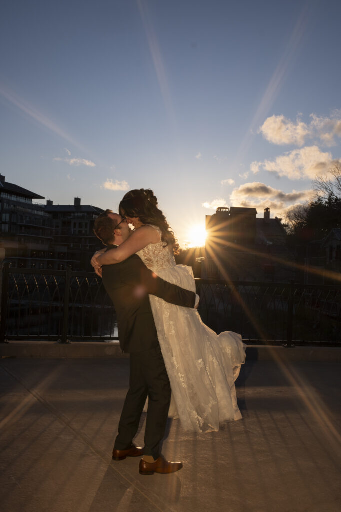 The groom lifts up his bride in an embrace at golden hour on the bridge near the Elora Mill in Elora, Ontario. 