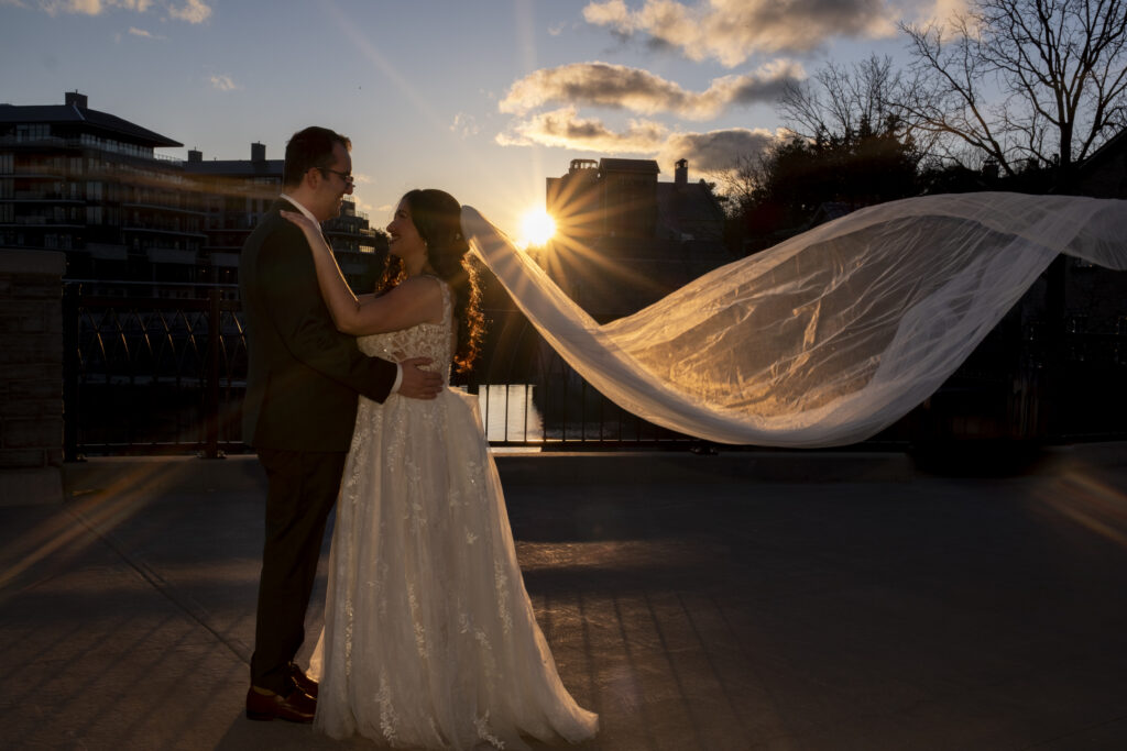 The bride's viel is caught in the breeze as she poses with her groom at golden hour on the bridge near the Elora Mill in Elora, Ontario. 